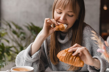 Young blonde woman with bang eating croissants at a cafe. Girl bite piece of croissant look joyful...