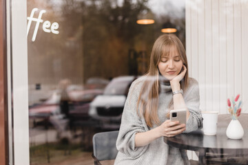 Young blonde woman with bang using phone sits in cafe at table hold smartphone, answering texts, phone call, letters, posting photos under windows glass. Girl messaging, paying using phone.