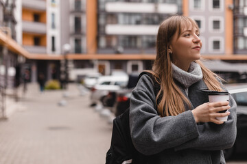 Cheerful young blonde woman with bang in casual wear drinking takeaway coffee while walking...