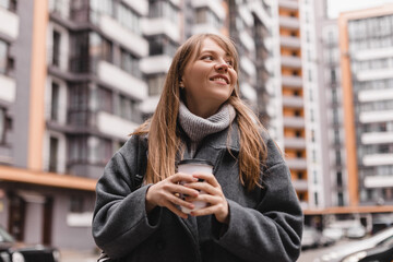 Cheerful young blonde woman with bang in casual wear drinking takeaway coffee while walking outdoors at street. Girl wear grey sweater, coat and black backpack. Woman walk or go home in courtyard.