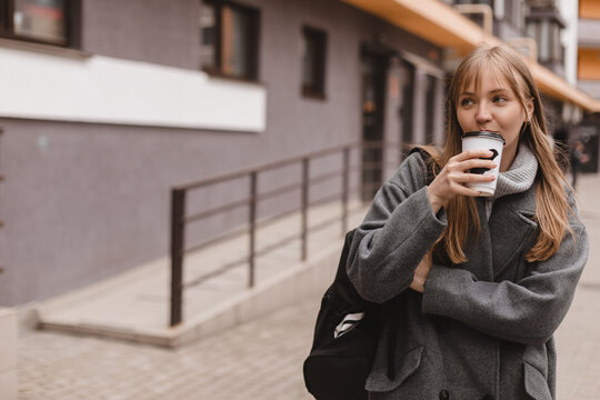 Cheerful young blonde woman with bang in casual wear drinking takeaway coffee while walking outdoors at street. Girl wear grey sweater, coat and black backpack. Woman walk or go home in courtyard.