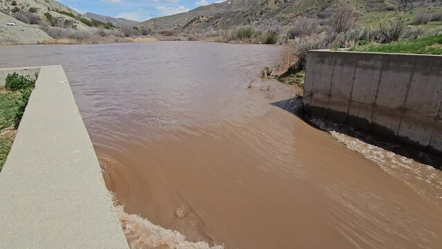 Muddy brown runoff water from mid-stream catch basin or retention pond overflows into spillway as record snow pack levels melt and threaten flooding.