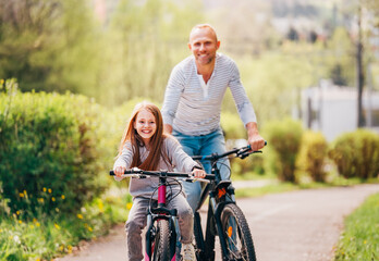 Fototapeta na wymiar Portraits Smiling father with daughter during summer outdoor bicycle riding. They enjoy togetherness in the summer city park. Happy parenthood and childhood or active sport life concept image.