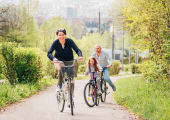 Obraz premium Smiling father and mother with daughter during summer outdoor bicycle riding. They enjoy togetherness in the summer city park. Happy parenthood and childhood or active sport life concept image.