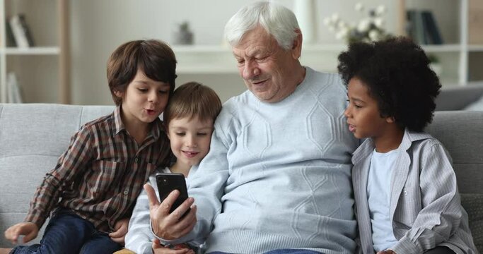 Cheerful curious diverse kids and senior granddad talking on online video call on mobile phone, resting close on sofa, using smartphone for Internet family communication, laughing