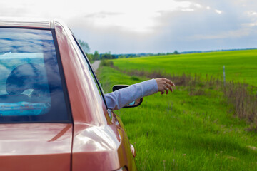 A woman's hand in a car window against the background of a green meadow and a stormy sky with sun rays. Beautiful summer scene with a car and beautiful vibrant nature. Summer and freedom.