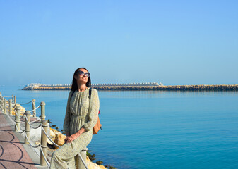 Fototapeta premium Young happy woman in a cute dress sits on a pier by the sea enjoying summer sunbeams