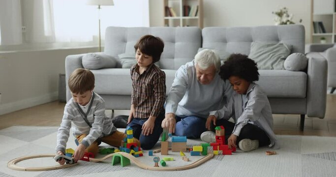 Senior grandpa watching diverse little grandkids playing construction learning game on heating floor, wheeling toy van, train on rail road, building city together, enjoying teamwork