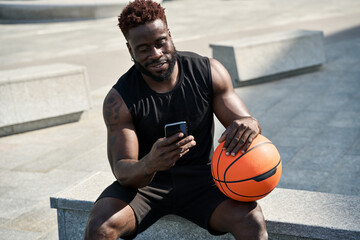 Fit sporty young African black ethnic man sitting outdoors holding basketball ball and mobile phone using apps, looking at smartphone, resting after street sport game with cellphone. - Powered by Adobe