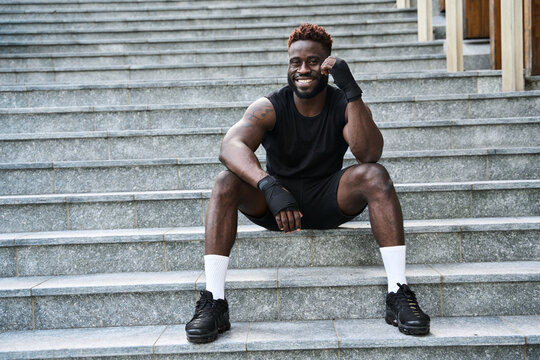 Fit happy sporty young black man boxer fighter sitting at urban stairs feeling confident. Strong African ethnic guy relaxing after street workout exercises or boxing, resting after training outside.