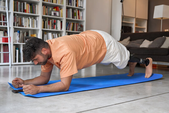 Sporty fit young man amputee with prosthetic leg disability prosthesis doing plank exercise on mat at home. Inclusive sport training for people with disabilities concept. Authentic shot