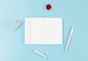 Empty notebook, menstrual cup, tampon and rose petal on a blue background.