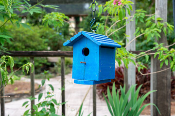 Fototapeta na wymiar A decorative bright blue single hole birdhouse on a fence. The birdhouse is attached to a wooden deck rail with green trees and shrubs in the background. There's a peg below the small hole. 
