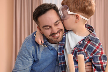 Son in protective glasses kissing father indoors. Repair work - obrazy, fototapety, plakaty