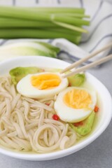 Bowl of delicious rice noodle soup with celery and egg on light grey table, closeup