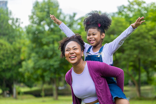 African American mother is playing piggyback riding with her young daughter while having a summer picnic in the public park for wellbeing and happiness concept