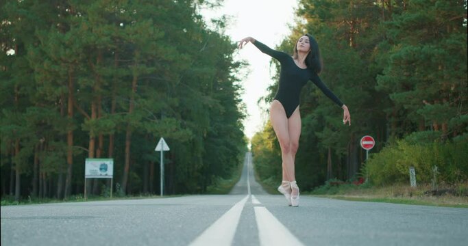 Flexible young girl dancer performing Classical ballet dance in forest on road on summer day. attractive caucasian lady in black bodysuit is flexible graceful. elegancy, dance, art, ballet