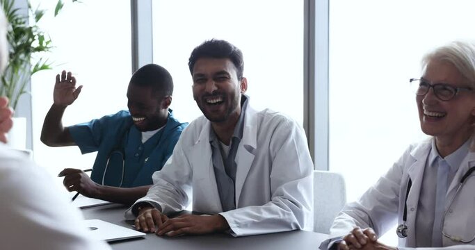 Laughing team of happy medical workers having friendly, nice conversation at group meeting in conference room in clinic. Multinational young and mature GP professionals communicating during seminar