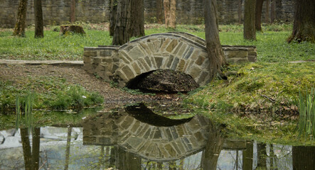Kamienny mostek pośród drzew z widocznym odbiciem z wodzie. Wokół budząca się do życia zieleń.
