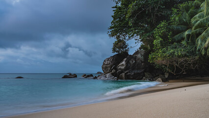 The waves of the turquoise ocean are foaming on the sandy beach. Boulders are scattered in the water. Coastal cliffs and tropical vegetation against a cloudy sky. Seychelles. Mahe. Long exposure