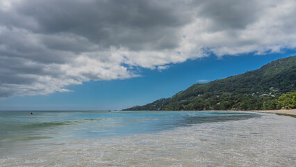 Fototapeta na wymiar The waves of the turquoise ocean spread over the sandy beach. A hill overgrown with tropical vegetation, against a background of blue sky and clouds. Seychelles. Mahe. Beau Vallon