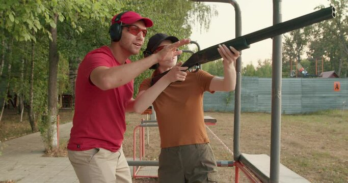 A Young woman with an instructor assisting with clay pigeon shooting. slow motion. Two adults in sunglasses and rifle vest practicing fire weapon shooting Experienced shooters aiming shotgun outdoors