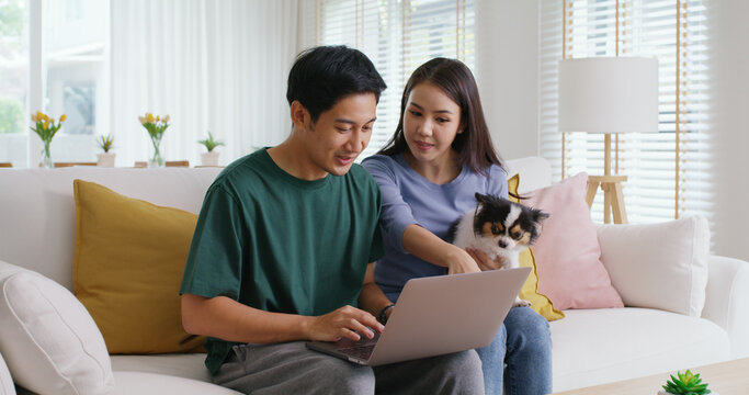 Asia people young family man woman smile happy sit at home sofa couch pay for hotel booking, search choose, buy ticket on laptop social media app travel trip getaway plan tour with cute small pet dog.