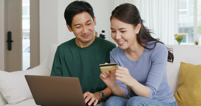 Secure pay gift card spend money buy ticket order from retail store on social media app. Asia people happy man woman, husband wife young couple sit at home sofa enjoy fun talk smile laugh relax.