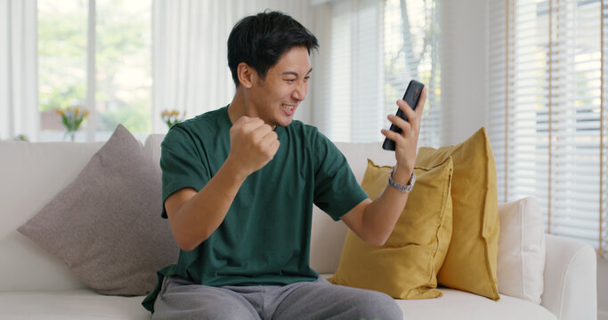 Asia young people teen man read good news message sitting at home sofa couch happy fun joy laugh smile in job hired result on mobile, award bonus, win online trade, sale order, loan money pay approve.