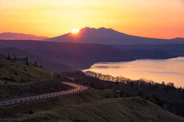 遠くの山から朝陽が昇る湖の脇の夜明けの峠。