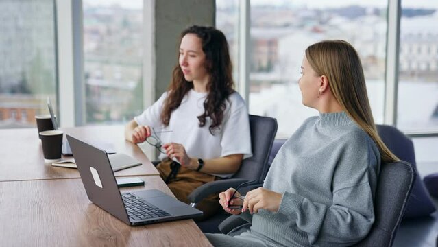 Two female colleagues take a break from work on laptops for a moment. Hard-working employees put on glasses and start typing again.