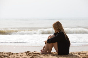 Beautiful young blonde woman in black shirt sits on the shore of the beach sad and depressed. The woman is pensive and looks at the ground exhausted and tired. The woman is suffering.
