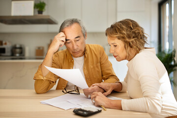Concentrated senior spouses sitting at table full of papers and counting monthly expenses, kitchen...