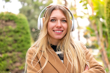 Young pretty blonde woman listening music with headphones at outdoors keeping the arms crossed in...