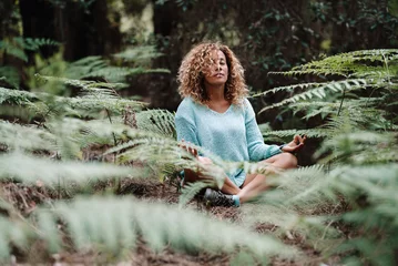 Tuinposter Health natural lifestyle meditation woman in the forest park with green tropical leaves all around. One healthy female people in yoga lotus position sitting on the ground in the woods. Wellbeing © simona