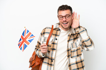 Young hispanic man holding an United Kingdom flag isolated on white background listening to something by putting hand on the ear