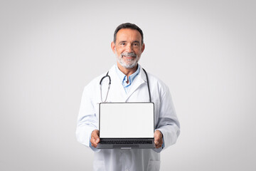 Happy european senior man doctor in white coat with stethoscope shows laptop with blank screen - Powered by Adobe