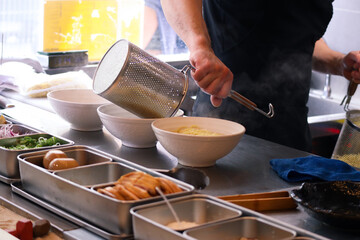 Bowl of Japanese ramen with hands putting the noodles inside it with ramen boiler on a countertop. With all ingredients to preparing Japanese ramen to serve in a restaurant 