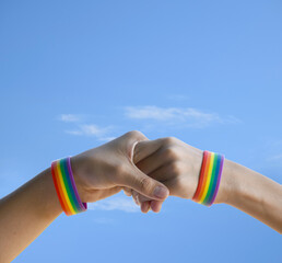 Closeup image of gay couple hands which wear rainbow wristband around on bluesky background with copy space.