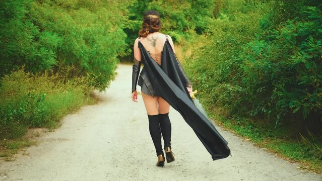 Fantasy woman warrior holds weapon sword in hands walks along road back rear view. Costume black leather clothes long road cloak flies in wind. Sexy girl warlike princess. summer nature green trees 4k
