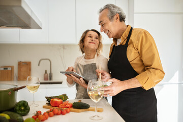 Senior caucasian couple preparing healthy dinner together, using digital tablet in cozy kitchen interior, free space