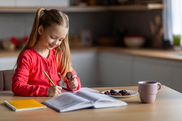 Fototapeta na wymiar Cute Little Girl Doing School Homework And Eating Cookies At Home