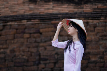 A pretty Vietnamese young Asian girl wearing beautiful Ao Dai a traditional Vietnam dress national costume color pink and a hat with an old brick wall. Ao Dai is popular in Vietnam.