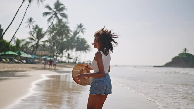 Multiethnic black woman runs on ocean beach on tropical vacation at exotic island. African american female turns, flirts running on sea shore with straw hat. Bipoc has fun on travel tour. Follow me.