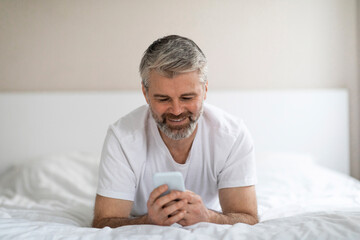 Relaxed happy mature man chilling in bed, using cell phone