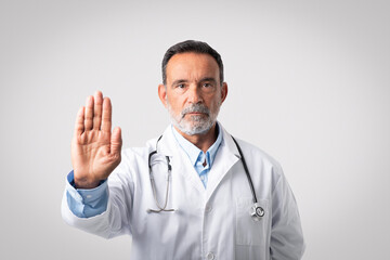 Serious confident european senior man doctor in white coat with stethoscope shows stop sign with...