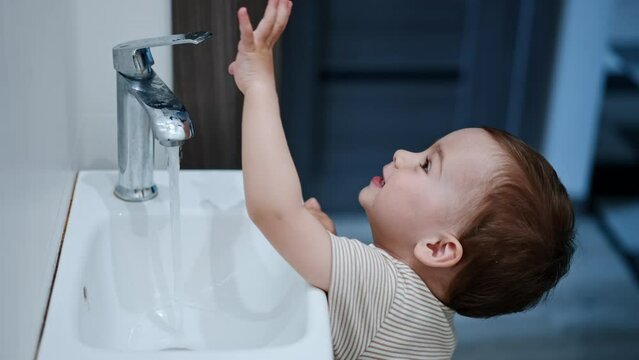 Curious little Caucasian toddler plays with water. Kid opens the faucet letting water flow.