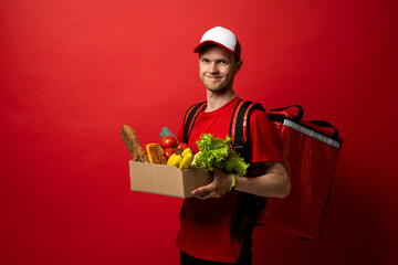 Delivery male employee in a red uniform holds paper cardboard package with organic groceries, fruits on a red background. Products delivery from shop or restaurant to home. Courier service.