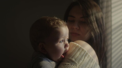 The child is in her mother's arms, yawns widely and looks with blue eyes. Sleepy baby. Millennial mother holds her son in her arms by the window of the house. A child's face in the sun.