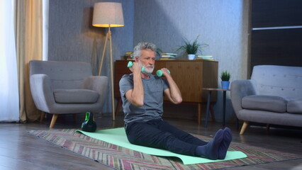 Portrait of a gray-haired senior man with a beard goes in for sports at home with dumbbells. In the living room exercises for pumping up the muscles of the arm beats. Old man in sportswear
